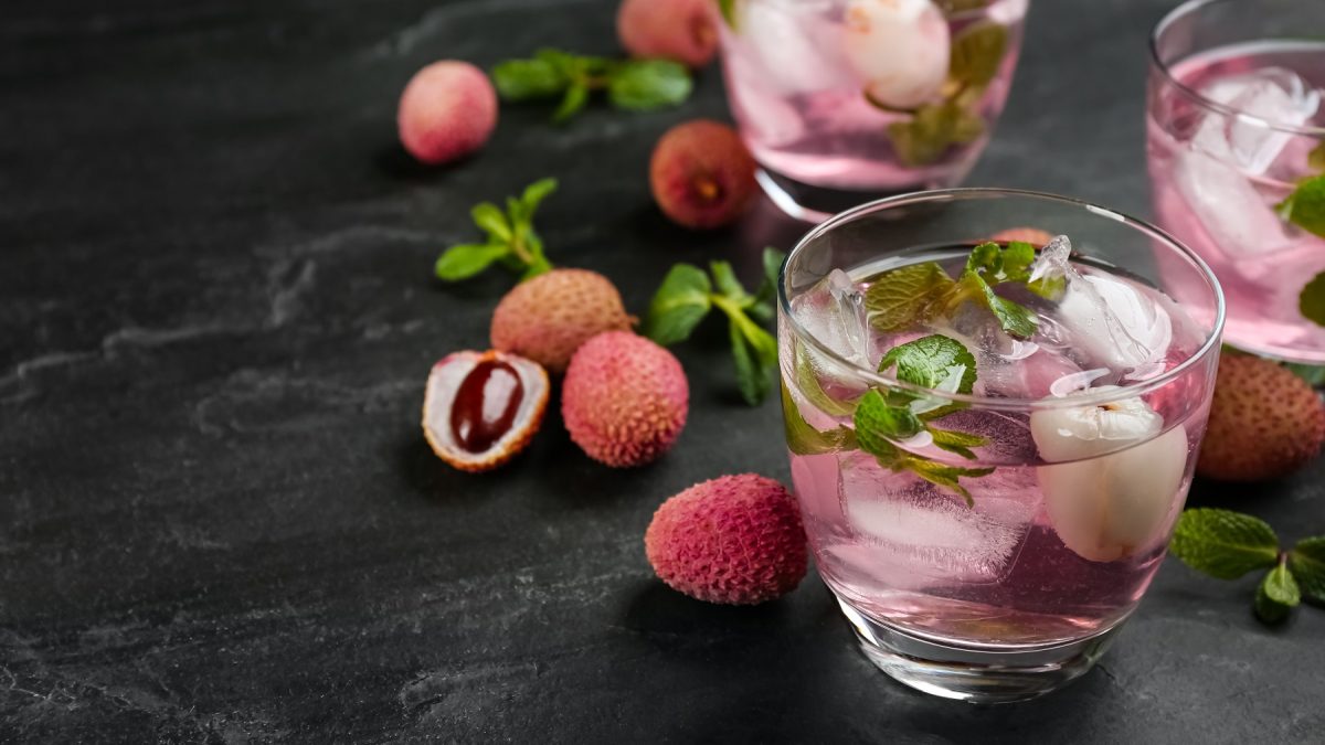 Drei Gläser mit Litschi Spritz auf dunkelm Untergrund, daneben frische Litschis in der Schale.