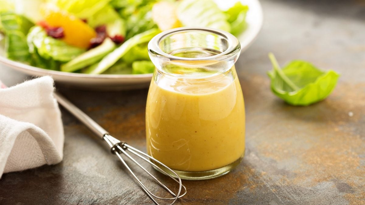 Ein Glas mit dem Apfel-Maracuja-Dressing auf eienm rustikalen Holztisch. Dahinter ein weißer Teller mit Blattsalat.