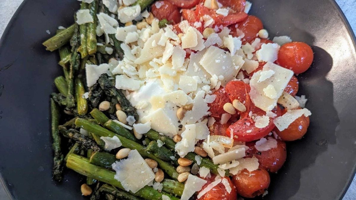 Gegrillter grüner Spargel und Kirschtomaten mit Pinienkernen und Parmesanspänen auf einem schwarzen Teller.
