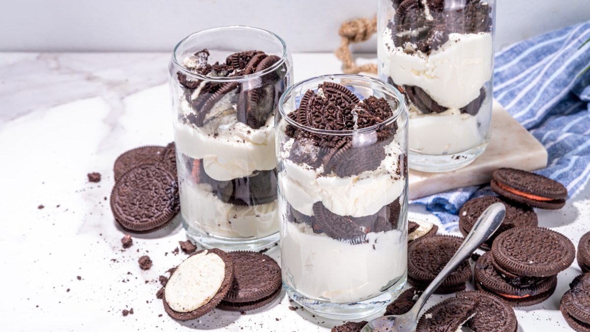 Drei Gläser mit dem Oreo-Trifle auf einem hellen Marmortisch mit blau-weiß-gestreifter Tischdecke.