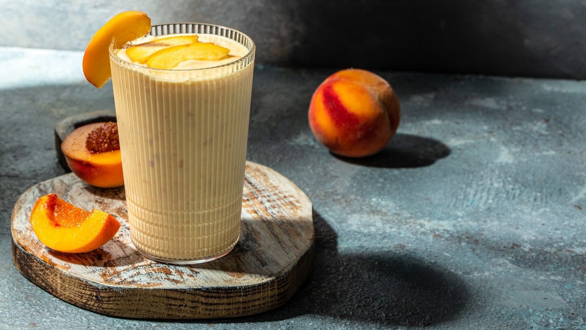 Ein Glas mit dem Shake auf einem Holzbrett und blau-grünlichem Marmorunter- und Hintergrund. Alles von der Seite fotografiert und drumherum frischer Pfirsich als Dekoration.