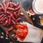Eine Auswahl an roten Mini-Würstchen und geräuchertem Lachs auf Holzbrett mit Pistazien, begleitet von zwei Bieren.