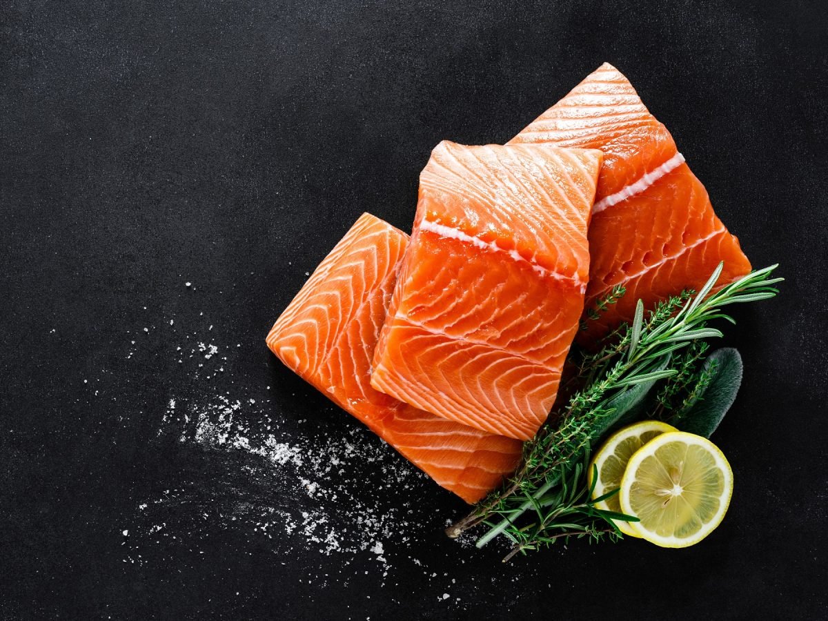 Drei Lachsfilets für Rezepte mit Lachs mit Zitronenscheiben und frischen Kräutern auf schwarzem Untergrund, leicht mit Salz bestreut.
