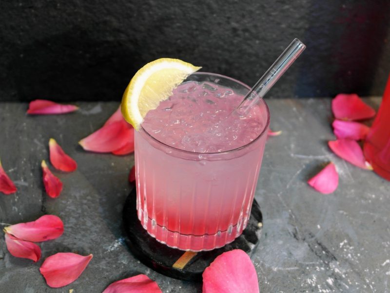 Ein Glas mit dem Sarti Lemon auf einem grauen Tisch mit pinken Blüten und einer Flasche Sarti. Alles vor schwarzem Hintergrund fotografiert.
