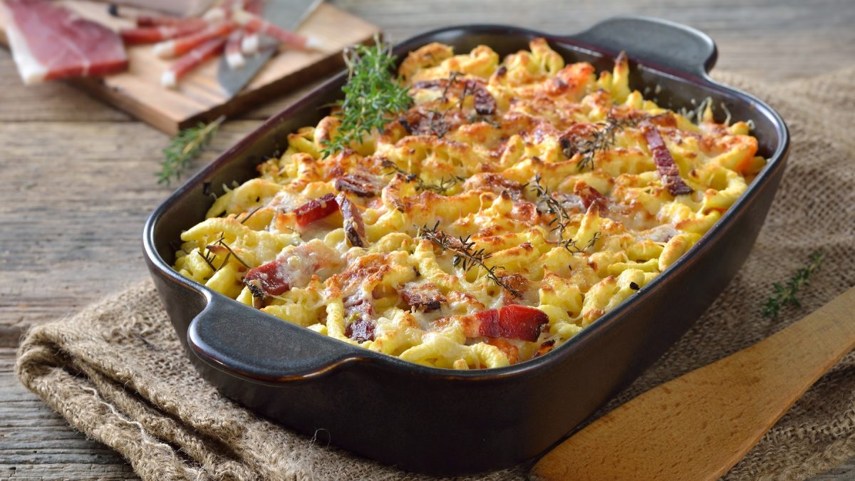 Eine schwarze Auflaufform mit dme Spätzle-Gratin auf einem rustikalen Holztisch. Im Hintergrund unscharf ganzer Speck zu erkennen. Alles von leicht schräg oben fotografiert.