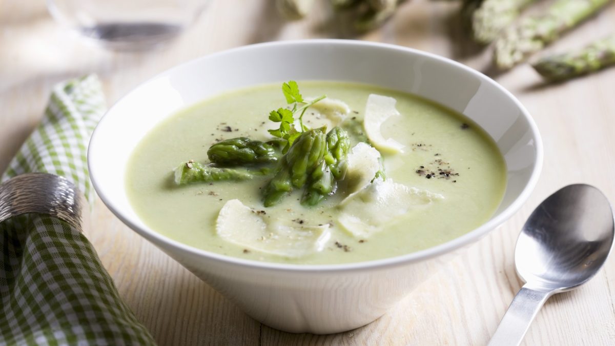 Spargelcremesuppe mit Parmesan in weißer Schüssel vor hellem Stoff-Hintergrund, daneben ein Löffel.