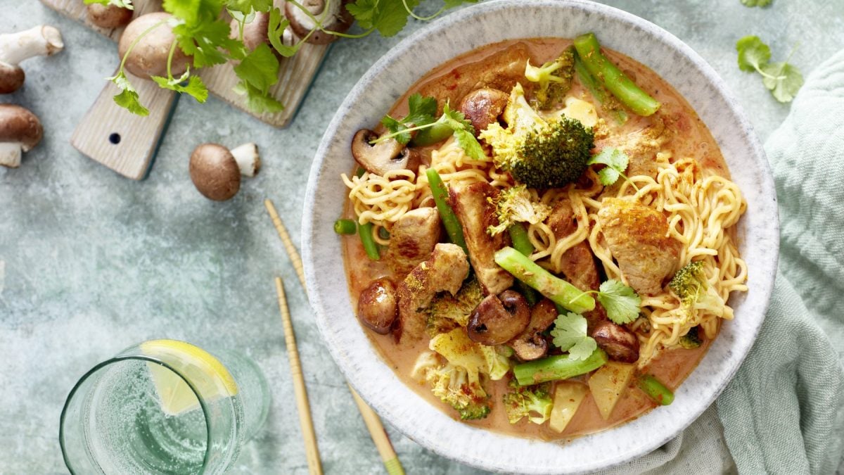 Thai-Curry mit Nudeln in einer weißen Schüssel angerichtet. Der Hintergrund ist grau und darauf sind Koriander und Champignons verteilt. Links unten auf dem Foto ein Glas gefüllt mit Wasser.