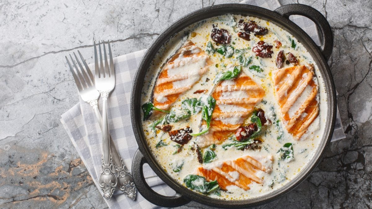 Eine Pfanne mit gegrilltem Lachs in cremiger Sauce mit Spinat und getrockneten Tomaten, auf grauem Marmorhintergrund, zwei silberne Gabeln daneben.