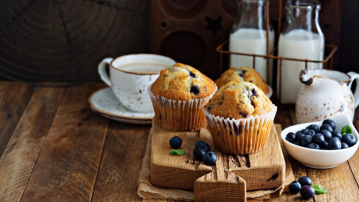Zwei vegane Heidelbeer-Muffins auf einem Holzbrett, daneben ein Becher Kaffee, Blaubeeren in einer Schale und Milchflaschen im Hintergrund.