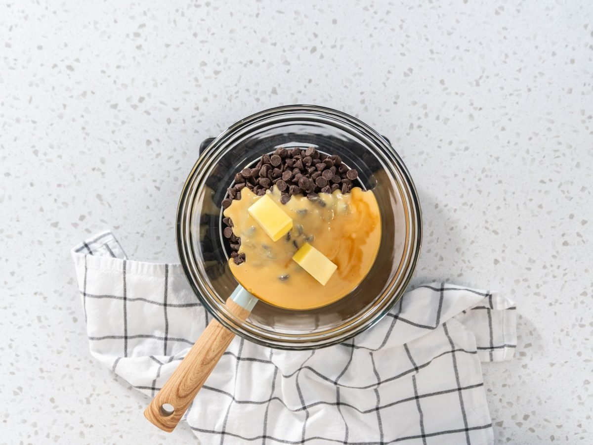 Glasschüssel für Wasserbad mit Schokoladenstückchen, Butter und Karamellsauce auf kariertem Küchentuch auf hellem Küchenarbeitsplatte.
