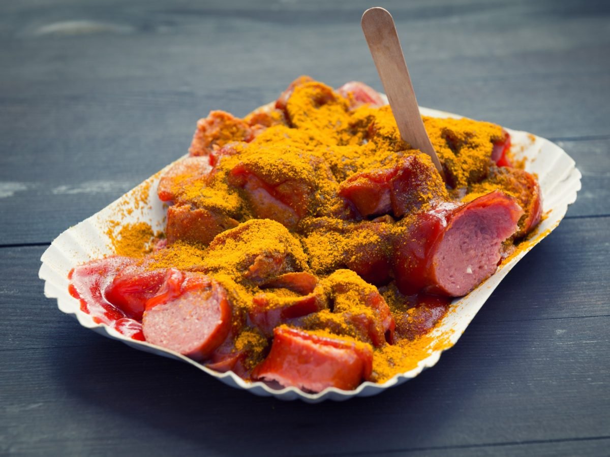 Eine weiße Pappschale mit roter Currywurst und Sauce auf einem dunklen Holztisch.
