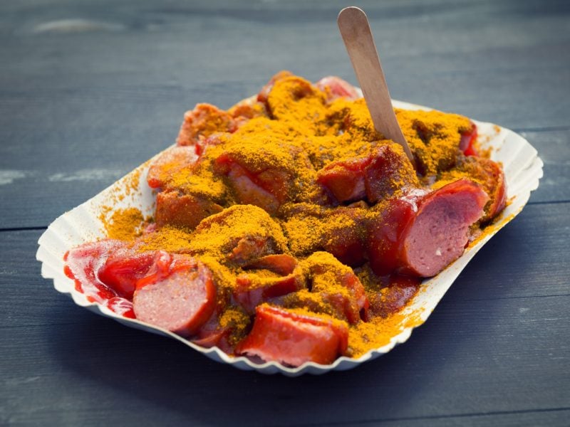 Eine weiße Pappschale mit roter Currywurst und Sauce auf einem dunklen Holztisch.
