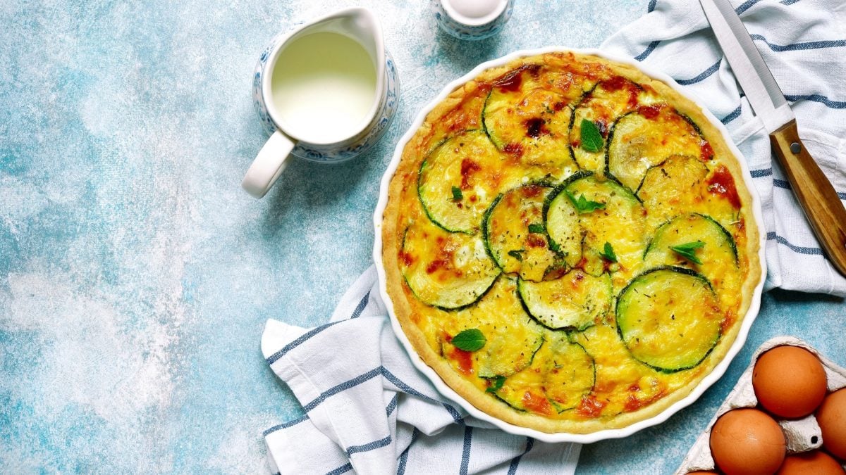 Draufsicht: Eine runde, weiß Form mit einer Zucchini-Quiche mit Käse auf hellblauem Untergrund. Ebenfalls zu sehen sind frische Eier, ein Küchenmesser, ein Kännchen mit Sahne und ein Küchentuch.