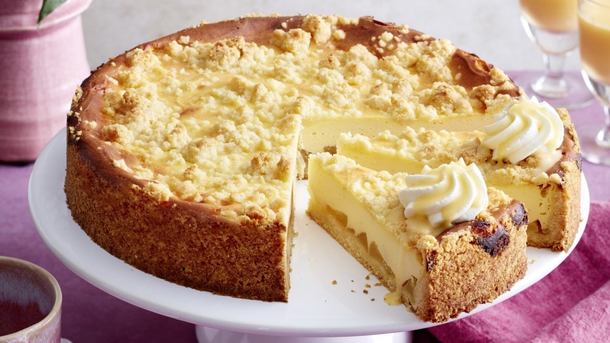 Angeschnittener Apfel-Käsekuchen mit Streuseln und Sahne auf Servierplatte.