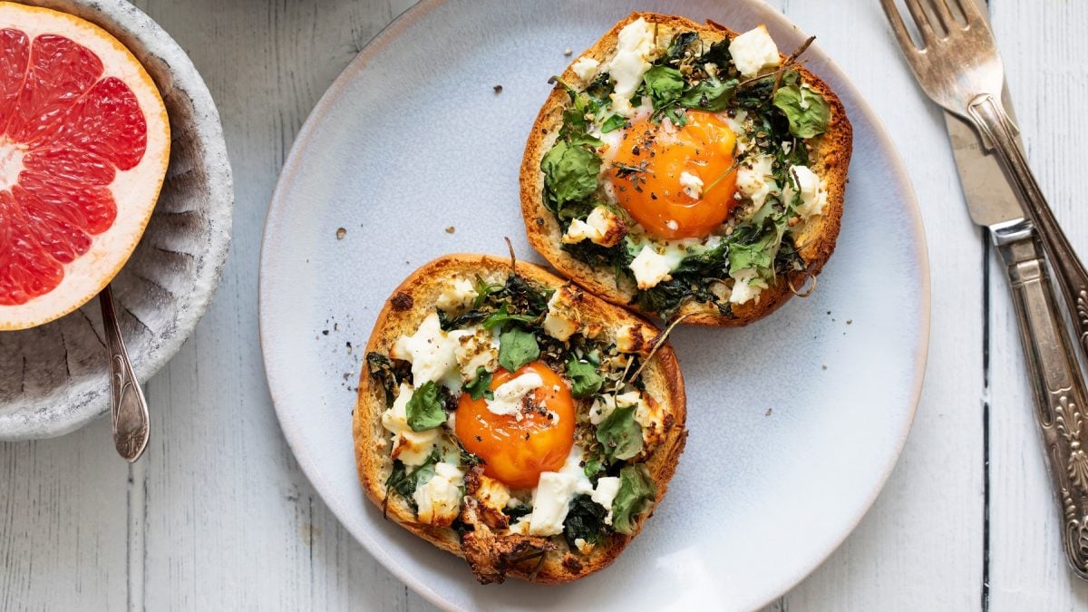 Zwei belegte Brothälften mit Spinat, Feta und Spiegelei auf einem hellblauen Teller, daneben eine halbe Grapefruit.