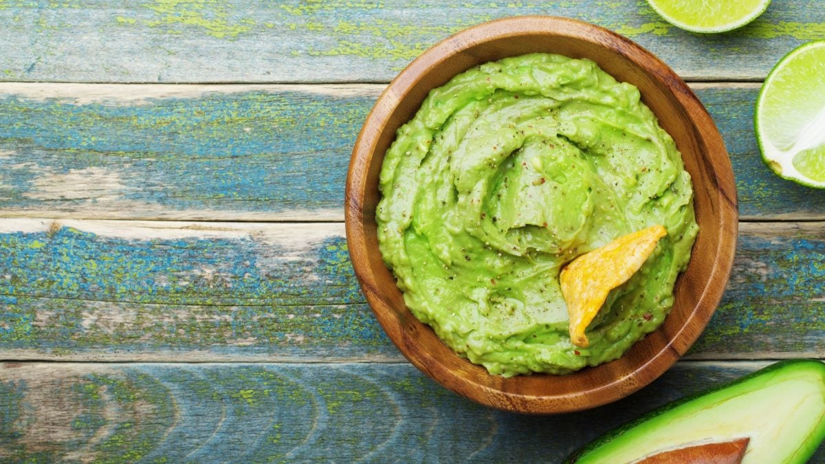 Draufsicht: Eine blauer-mediterraner Untergrund aus Holz. Auf dem steht eine Schüssel mit einem Avocado-Dip mit Limette. Daneben liegen eine halbe Avocado und zwei Limetten-Schnitzer,