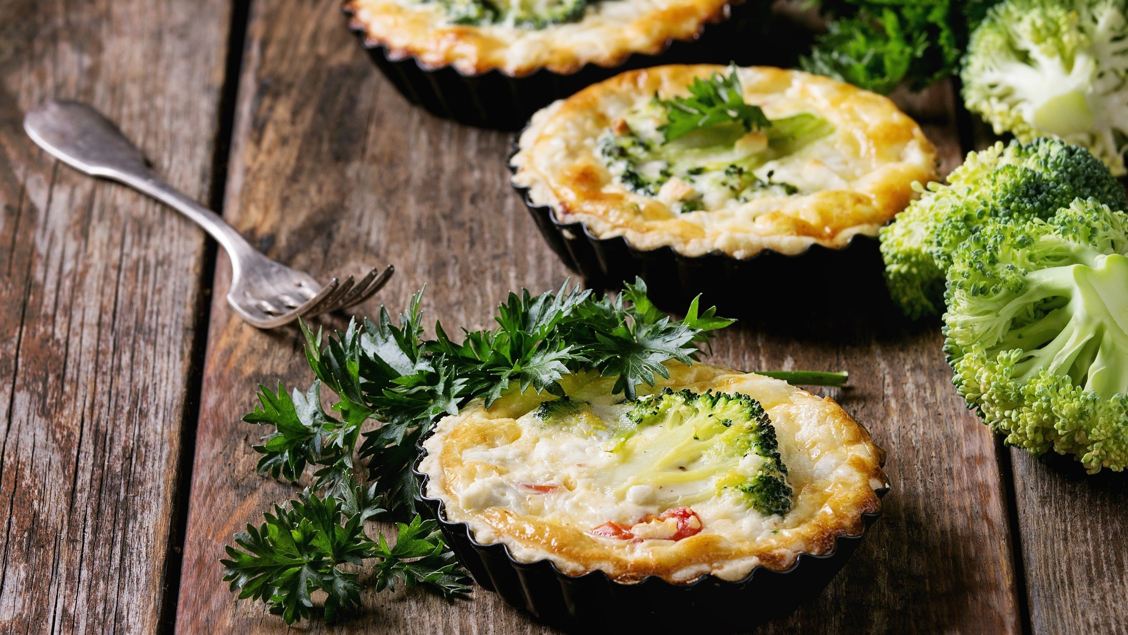 Draufsicht, schräg: Drei Brokkoli-Quiche in kleinen Tarte-Formen auf einem rustikalen Holztisch.