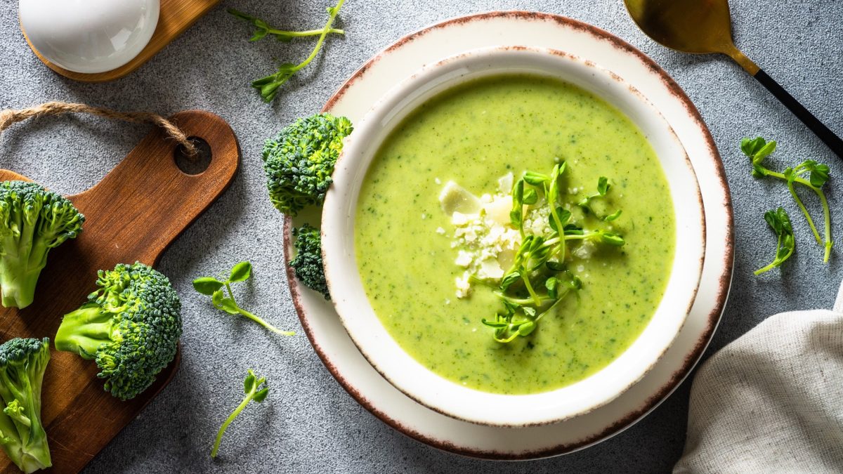 Draufsicht: Auf einem bläulichem Untergund steht eine runde Schale auf einem Teller. In der Schüssel ist eine Brokkoli-Suppe mit Kartoffeln. Drum herum liegen Brokkoli-Röschen und Brokkoli-Sprossen sowie ein Löffel.