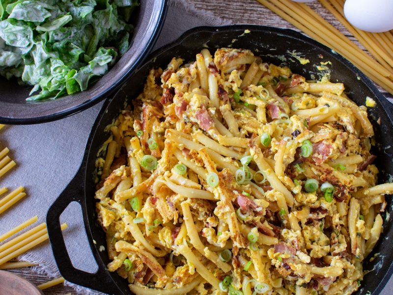 Draufsicht: Eine Pfanne Schinken-Nudeln mit Frühlingszwiebeln. Drum herum liegen rohe Nudeln und ein rohes Ei. Daneben steht eine Schüssel mit einem frischen grünen Salat.