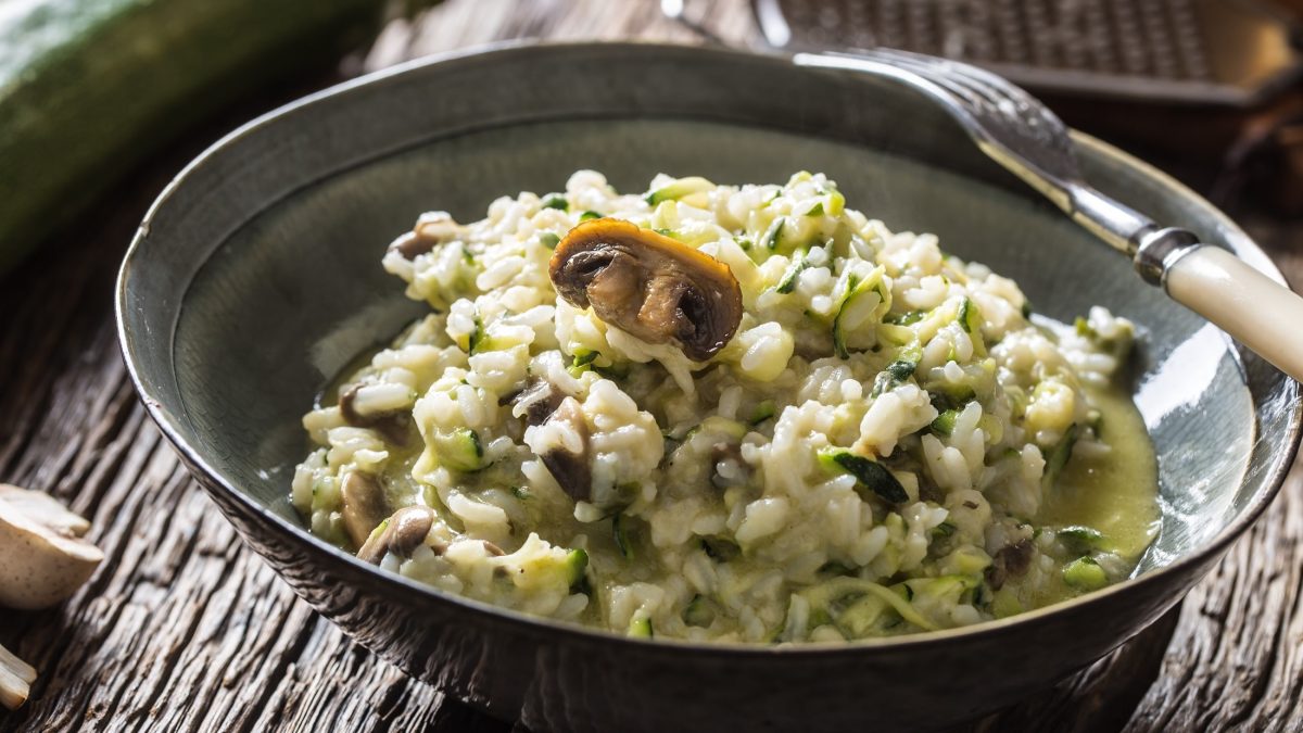 Draufsicht, schräg: Eine weiße Schüssel Zucchini-Risotto mit Pilzen auf einem rustikalen Untergrund aus Holz.