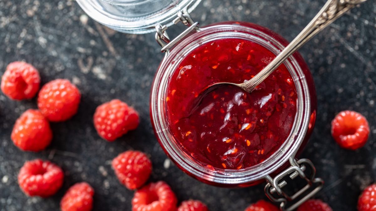 Ein Glas mit Himbeermarmelade, daneben verstreute frische Himbeeren auf einem dunklen Steinuntergrund.