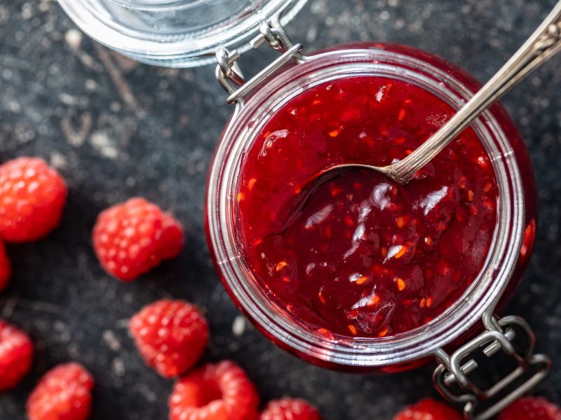 Ein Glas mit Himbeermarmelade, daneben verstreute frische Himbeeren auf einem dunklen Steinuntergrund.