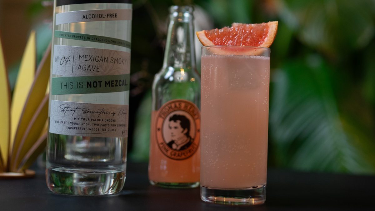 Paloma Undone in einem Highballglass mit Grapefruitschnitz, daneben Flaschen von Undone und Thomas Henry.