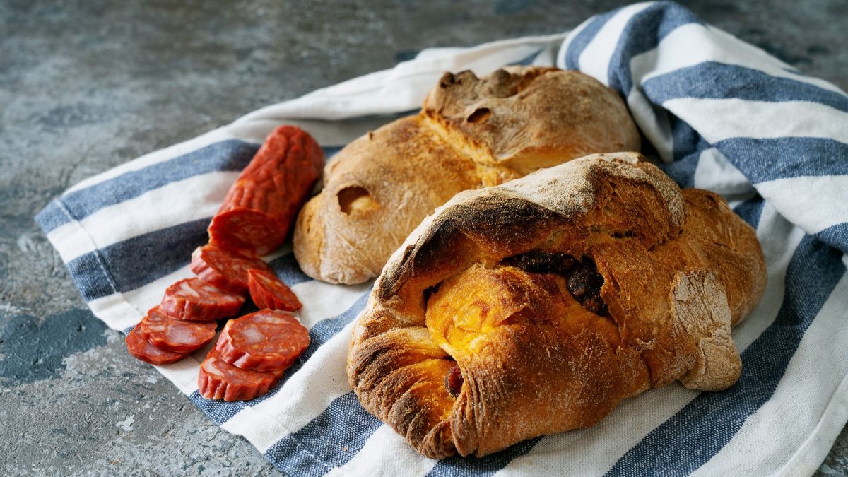 Zwei rustikale Pão com Chouriço und aufgeschnittene Chouriço liegen auf einem blau-weiß gestreiften Tuch auf grauem Steinboden.