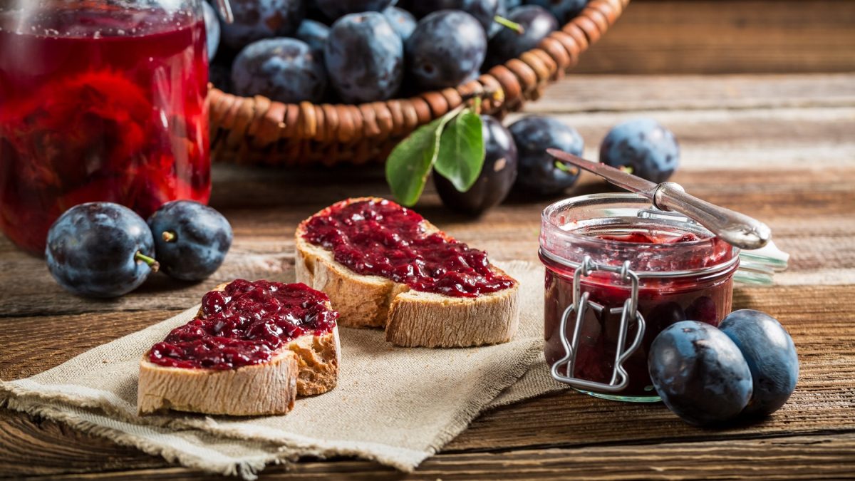 Zwei Scheiben Brot mit Pflaumenmarmelade, daneben ein Glas Marmelade und frische Pflaumen auf rustikalem Holztisch.