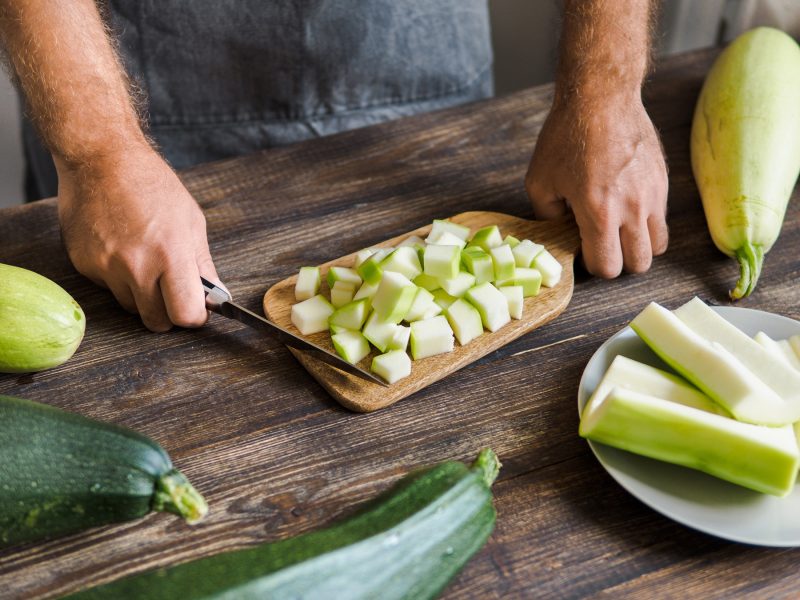 Ein Mann in grauer Schürze würfelt helle Zucchini auf einem Holztisch, daneben ganze und geschnittene Zucchini.