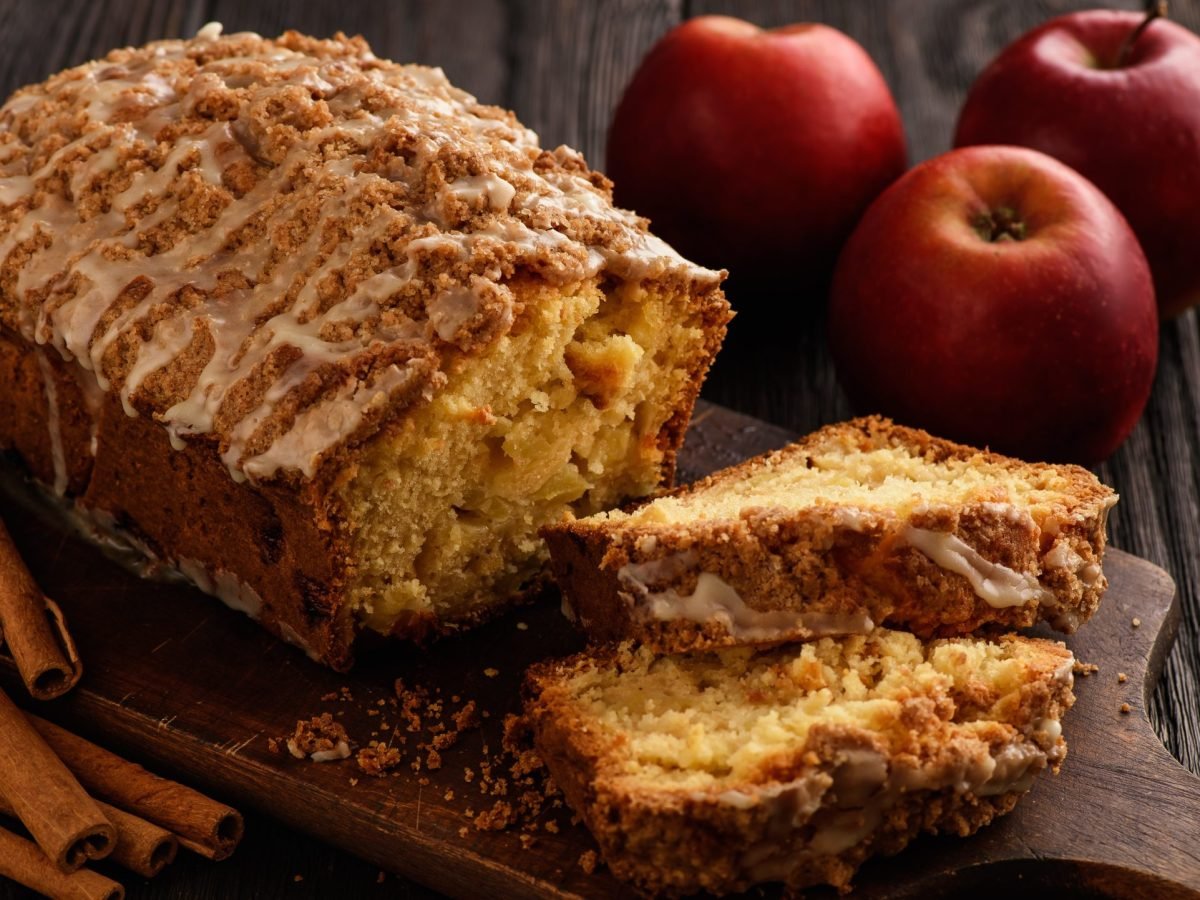 Apfel-Zimt-Kuchen auf einem Holzbrett auf hölzernem Hintergrund. Rechts davon Äpfel und links davon Zimtstangen.