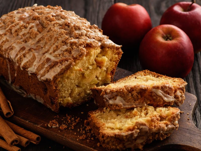 Apfel-Zimt-Kuchen auf einem Holzbrett auf hölzernem Hintergrund. Rechts davon Äpfel und links davon Zimtstangen.