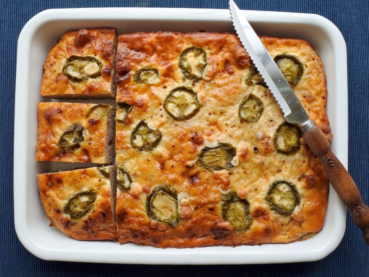 Eine Ofenform mit dem Maisbrot direkt von oben auf dunkelblauem, mattem Untergrund fotografiert. An der Seite liegt noch ein Messer mit Holzgriff.