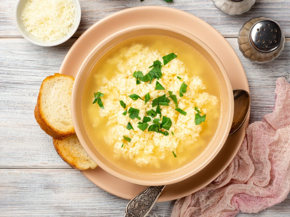 Draufsicht: Eine italienische Stracciatella-Suppe in einer Schale auf einem hellen Untergrund aus Holz. Drumherum liegen eine Serviette und Brotscheiben.