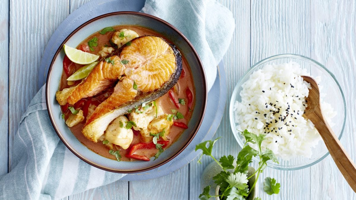 Lachskoteletts in Currysauce in einem tiefen Teller, daneben eine Schüssel mit Reis, Sesam und Petersilie auf hellblauem Holzuntergrund.