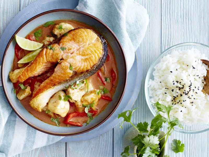 Lachskoteletts in Currysauce in einem tiefen Teller, daneben eine Schüssel mit Reis, Sesam und Petersilie auf hellblauem Holzuntergrund.