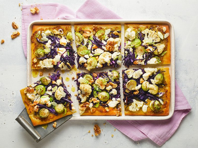 Blech mit rechteckiger Pizza, belegt mit Rosenkohl, Rotkohl, Blumenkohl, Walnüssen und Ziegenkäse auf einem rosafarbenen Tuch.