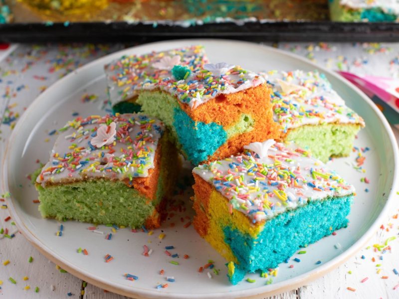 Bunte Kuchenstücke in Blau, Grün und Orange mit Zuckerguss und Streuseln auf weißem Teller, dekoriert mit bunten Servietten und verstreuten Streuseln – auch bekannt als Papageienkuchen.