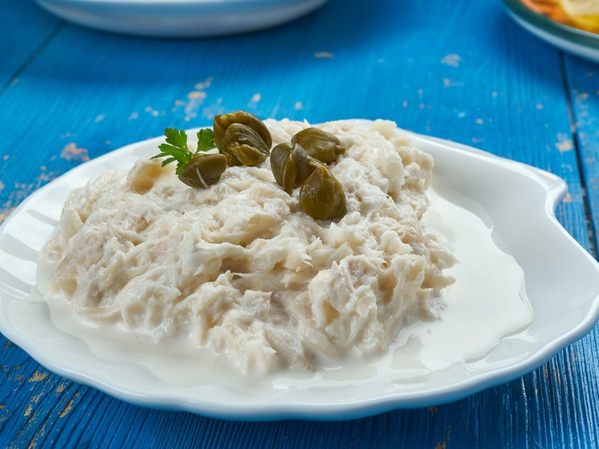 Baccalà mantecato mit Kapern auf einem weißen Teller. Der steht auf einem blauen Untergrund aus Holt. Die Anmutung ist sommerlich, mediterran.