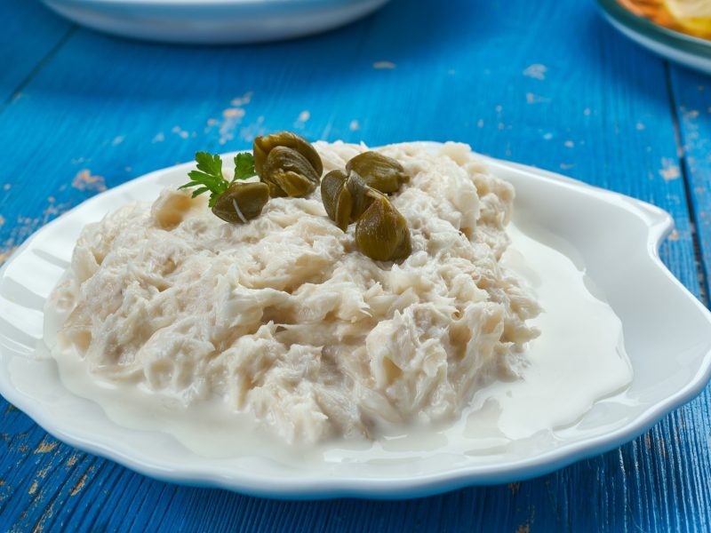 Baccalà mantecato mit Kapern auf einem weißen Teller. Der steht auf einem blauen Untergrund aus Holt. Die Anmutung ist sommerlich, mediterran.