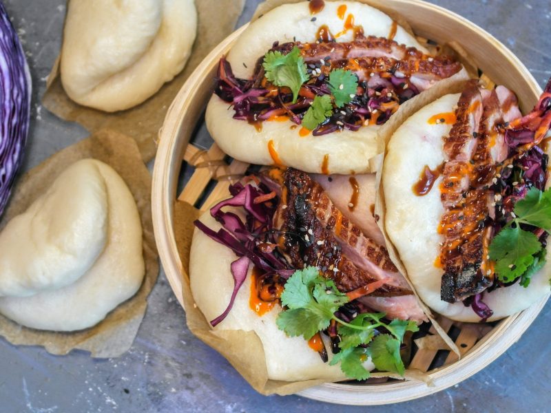 Ein Dampfkorb mit den Bao-Buns gefüllt auf einem grauen Steintisch. Daneben angeschnittener Rotkohl und ungefüllte Dampfbrötchen. Alles direkt von oben fotografiert.