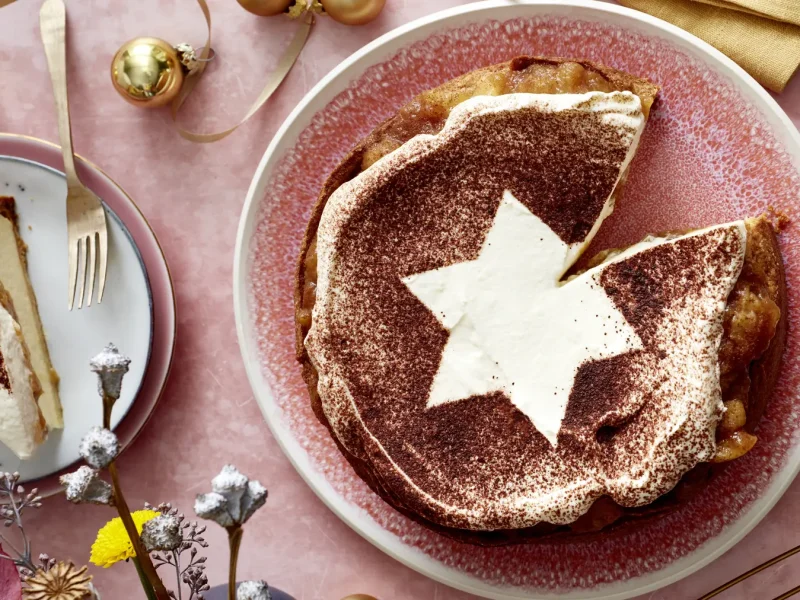 Bratapfel-Käsekuchen auf einem rosa Hintergrund. Links ein kleiner Teller mit einem Stück angerichtet und goldene Weihnachtskugeln verteilt.