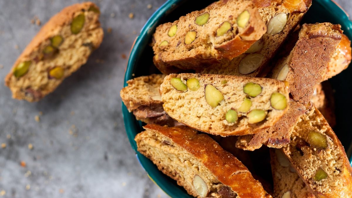 Draufsicht: Mehrere Cantuccini mit Pistazien liegen auf einem dunklen Teller. Ein Keks liegt neben dem Teller auf einem hellen Untergrund.