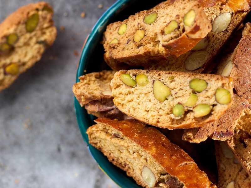 Draufsicht: Mehrere Cantuccini mit Pistazien liegen auf einem dunklen Teller. Ein Keks liegt neben dem Teller auf einem hellen Untergrund.