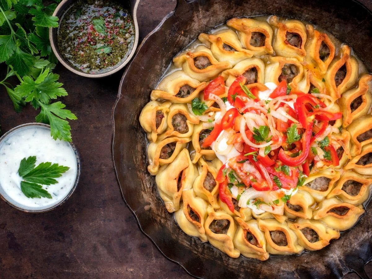 Manti mit Hackfleisch in einer ofenfesten gusseisernen runden Form. Die steht auf einem dunklen Untergrund. Drum herum ist eine Sauce aus Joghurt, eine Schale mit einer Sauce und frische Petersilie.