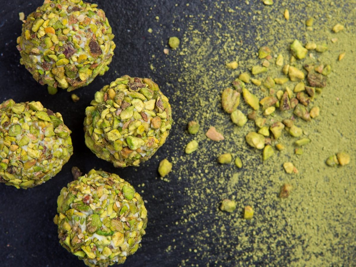 Pistazien-Energy-Balls auf einem schwarzen Untergrund. Drum herum sind klein gehackte Pistazien sowie ein grünes Pulver.