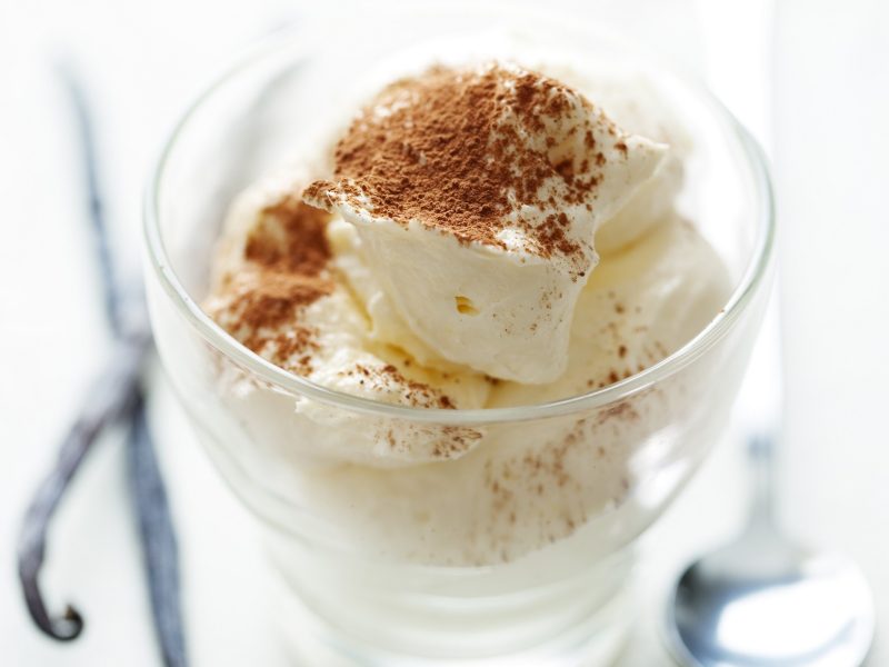Vanille-Mousse mit Kakaopulver bestreut in einem Dessertglas. Auf dem weißen Untergrund liegen drei schmale Vanilleschoten. Draufsicht, schräg.