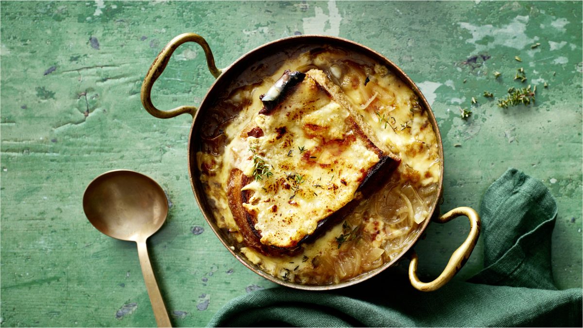 Draufsicht: Französische Zwiebelsuppe im Topf, daneben ein Löffel auf grünem Holzuntergrund.