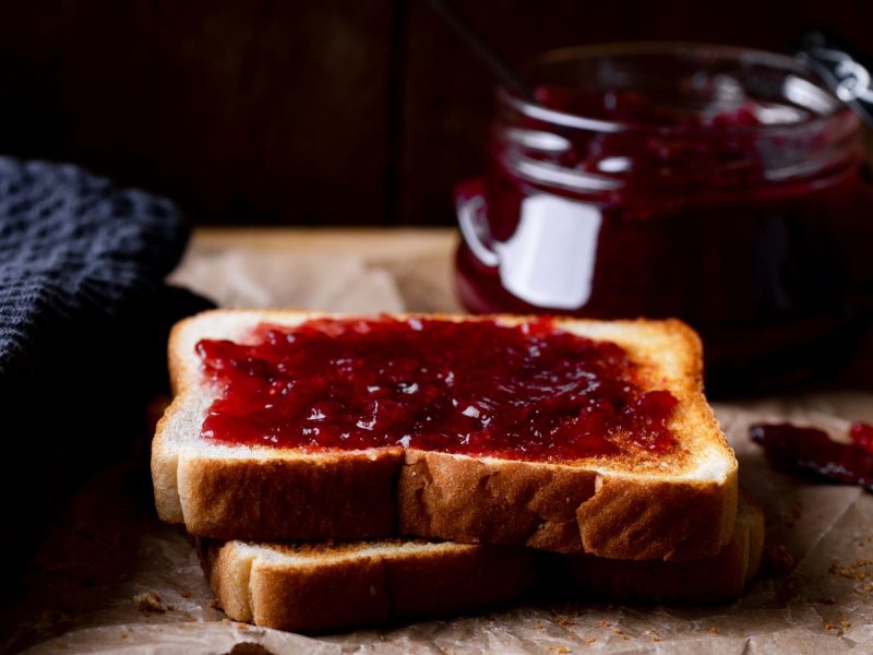 Zwei goldbraune Toastscheiben mit leuchtender Glühweinmarmelade, daneben ein Glas Konfitüre und ein Messer auf braunem Papier.