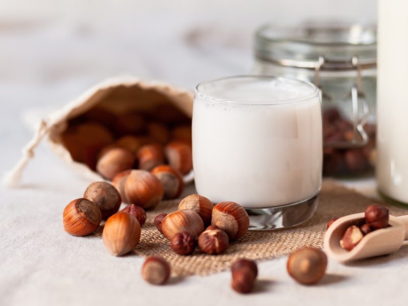 Glas mit Haselnussmilch von vorne fotografiert, daneben ungeschälte Haselnüsse sowie eine Flasche mit besagter Milch.